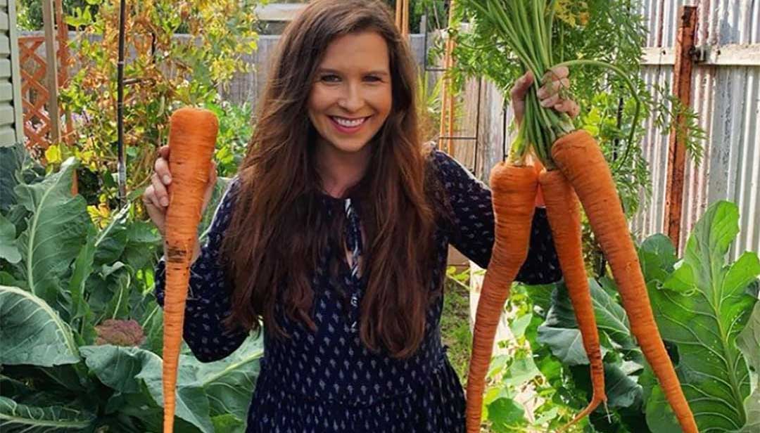 Woman Holding Up Giant Carrots As Part of the Grow It Local Wonky Veggies Competition