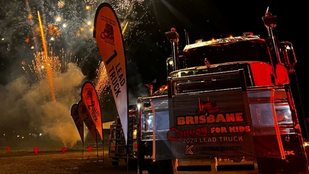 Nighttime Convoy Scene with a Large Red Lead Truck and Banners Reading Brisbane Convoy for Kids, with Fireworks in the Background.