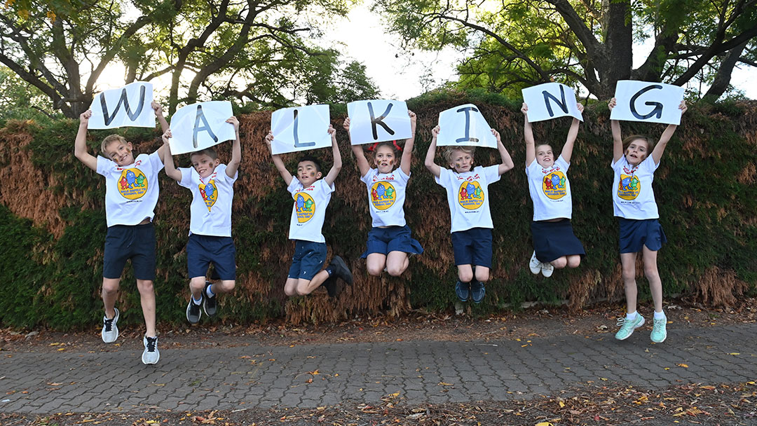 Eight children in white shirts leap in the air while holding up signs that spell 'WALKING' in front of a hedge to represent National Walk Safely to School Day