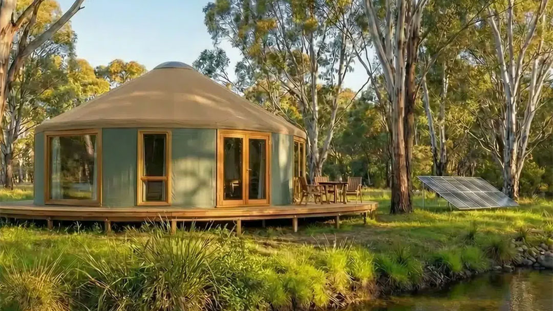 Yurt3 | Kids in Brisbane, Gold Coast, Sunshine Coast | Kotc Round Beige Yurt on a Wooden Deck Surrounded by Trees, with Outdoor Table and Chairs and a Nearby Solar Panel Array in a Sunny Forest Setting.