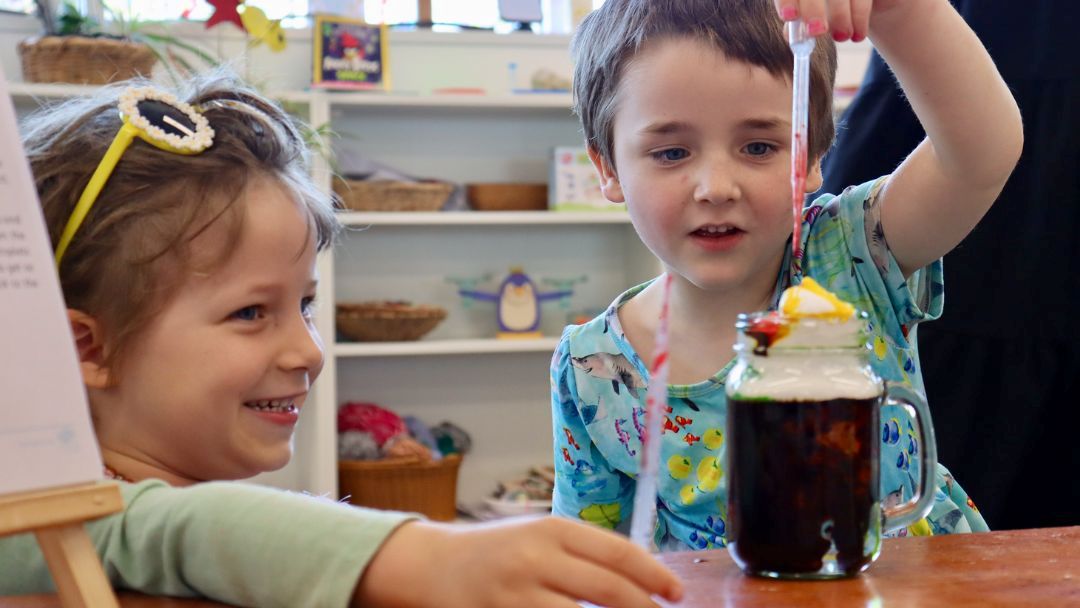 0514_compassindependentschool_hero | Kids in Brisbane, Gold Coast, Sunshine Coast | Kotc Two Smiling Children Enjoy a Festive Drink Activity, One Pouring a Striped Straw into a Mason Jar Filled with Dark Soda and Ice.