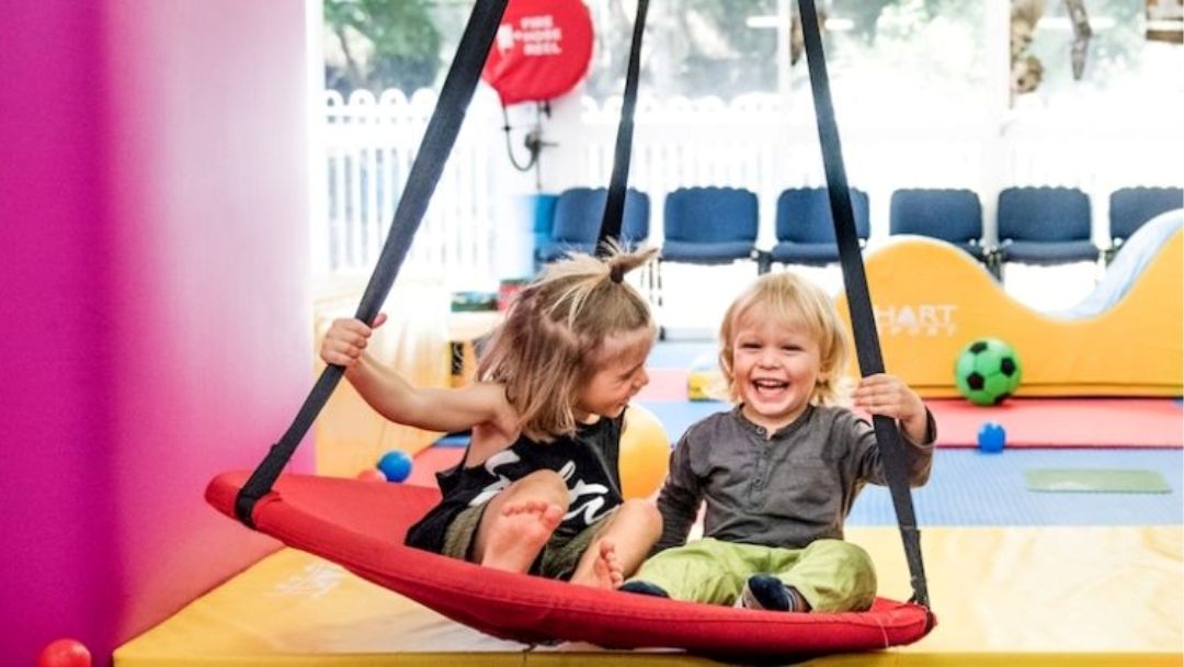 Two Smiling Toddlers on a Red Swing Inside a Colorful Indoor Play Area.