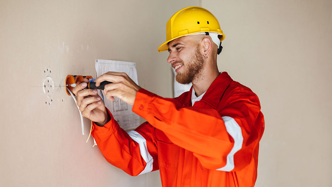 Construction Worker in a Bright Orange Safety Suit and Yellow Helmet Uses a Drill to Install Wiring in a Wall.