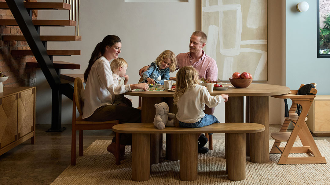 a Family of Four Sits Around a Round Wooden Table from Mocka Store in a Modern Dining Room, Sharing a Meal with a Fruit Bowl Nearby.