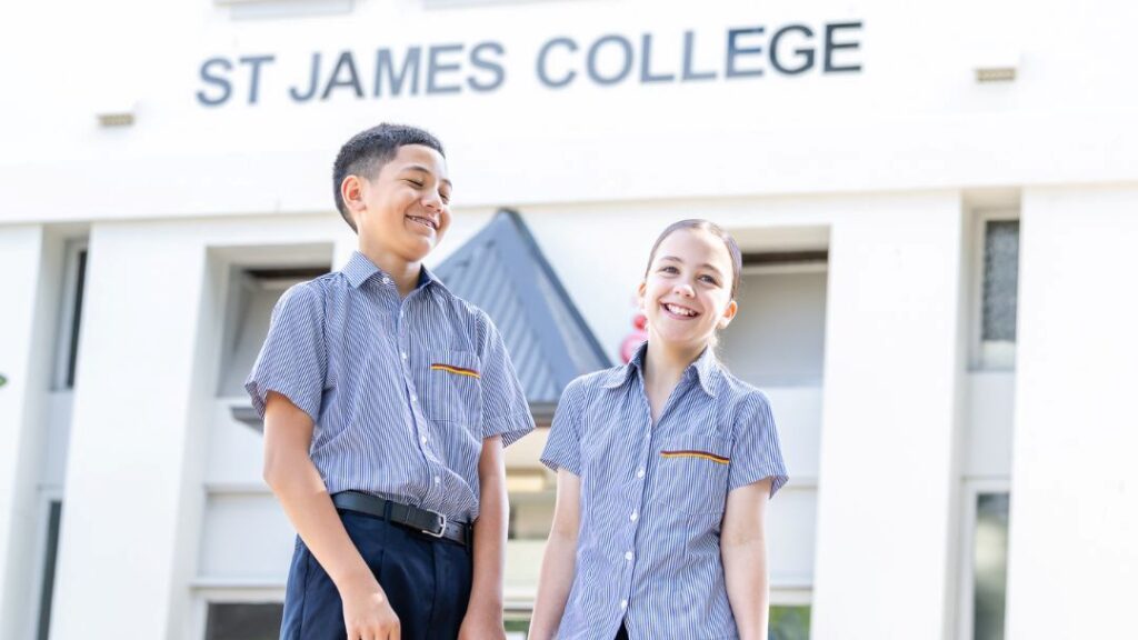 0601_stjamescollege_openday_hero | Kids in Brisbane, Gold Coast, Sunshine Coast | Kotc Two Smiling Students in Blue Striped Uniforms Stand Outside a White School Building with 'st James College' Signage Above.