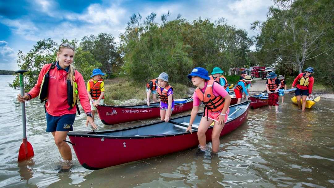 Group of children wearing life jackets preparing red canoes on a shallow shoreline during a canoeing trip.