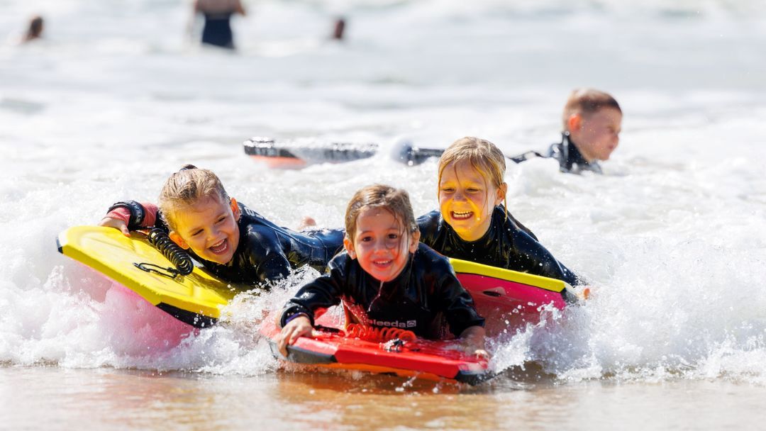 0609_departmentofsport_goldcoastrecreationprecinct_hero | Kids in Brisbane, Gold Coast, Sunshine Coast | Kotc Three Kids on Colorful Boogie Boards Ride a Small Wave at the Beach, Smiling and Wearing Wetsuits.