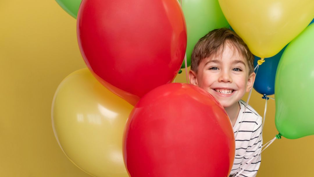 A smiling boy in a striped shirt among colorful birthday balloons (red, yellow, green) against a yellow wall.