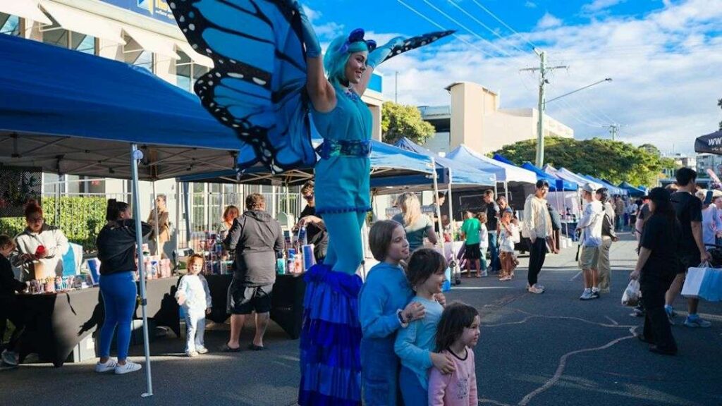 Blue Butterfly-costumed Performer on Stilts Entertaining Children at an Outdoor Market with Vendor Tents.