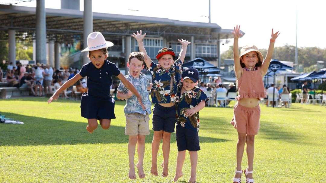 0611_sunshinecoastturfclub_schoolholidays_hero | Kids in Brisbane, Gold Coast, Sunshine Coast | Kotc Five Children Jump in the Air on a Sunny Park Lawn, Wearing Hats and Summer Outfits, with Spectators in the Background.