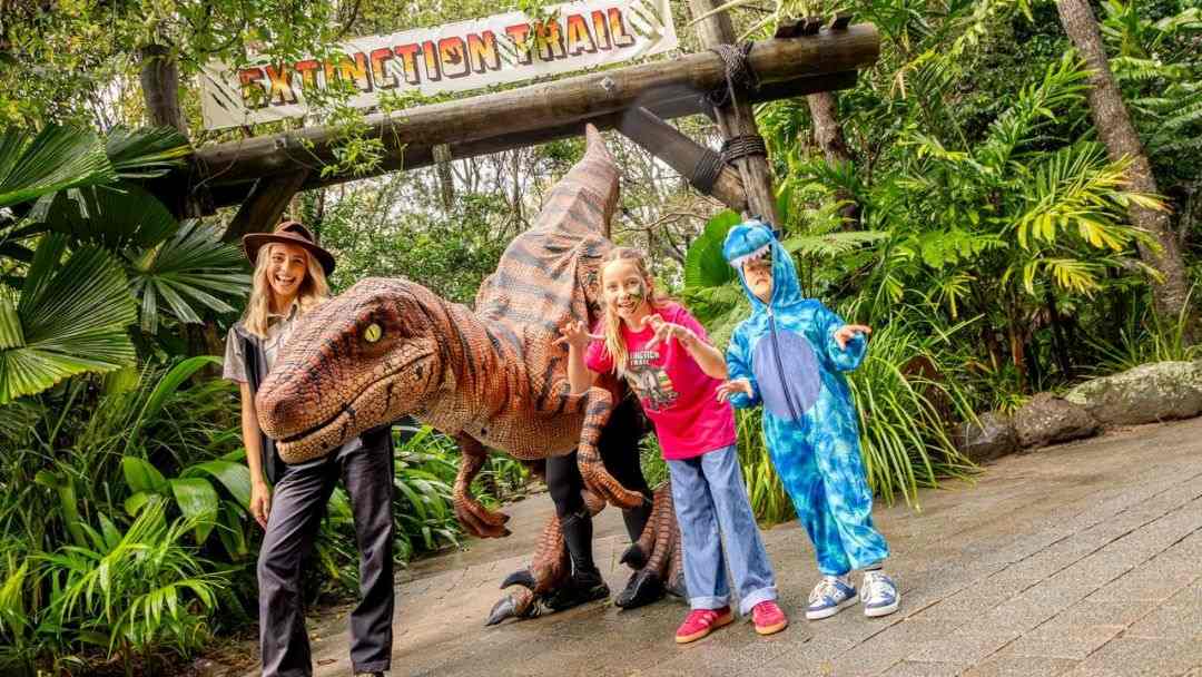People Pose with a Life-size Dinosaur Model Under a Sign Reading 'extinction Trail' in a Lush Outdoor Setting.