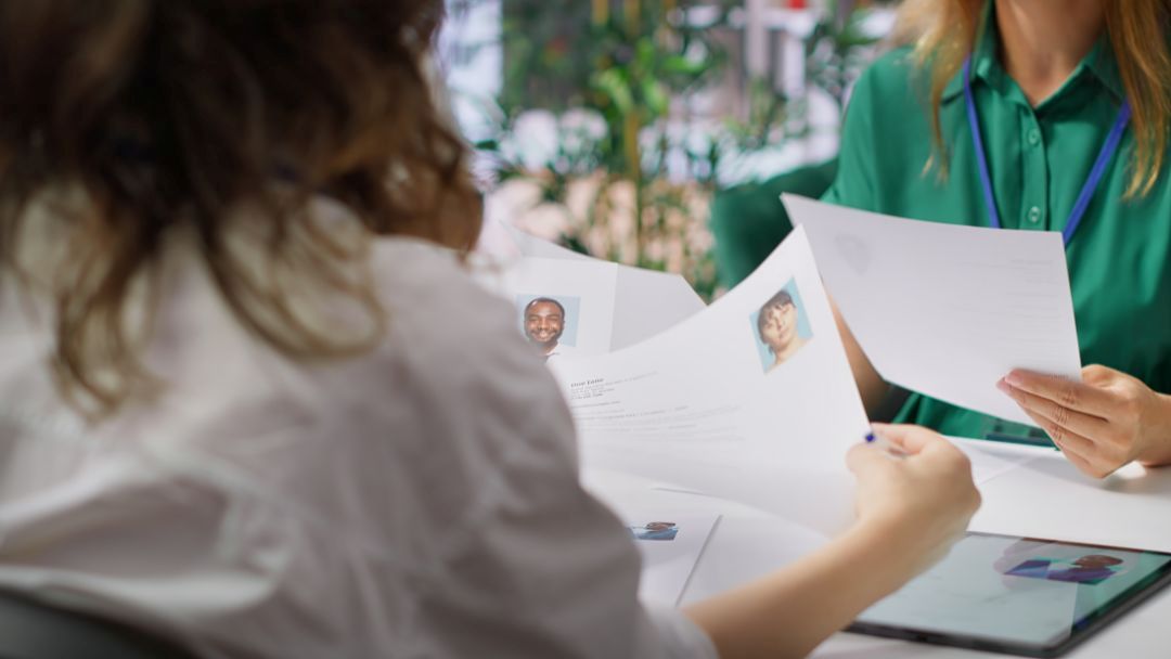 Two People Review Printed Resumes with Headshots at a Desk, a Tablet Visible on the Table