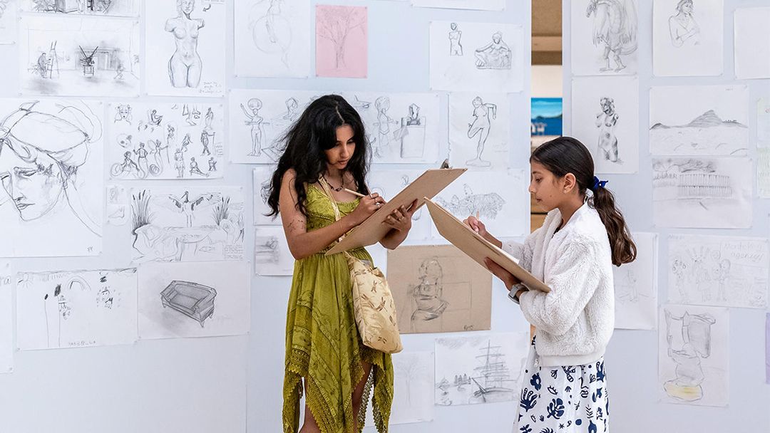 Two young girls studying sketches on clipboards in a studio with drawings taped to the wall behind them.