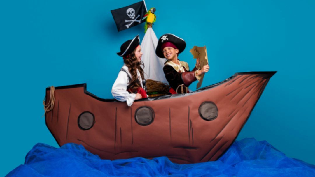 Two children dressed as pirates sit in a brown cardboard ship, a skull-and-bones flag above, against a blue background.