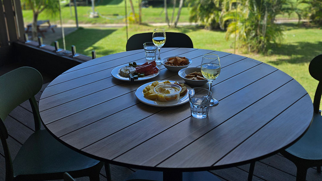 Round Wooden Patio Table Set with Plates of Appetizers and Glasses of White Wine, Overlooking a Grassy Garden.