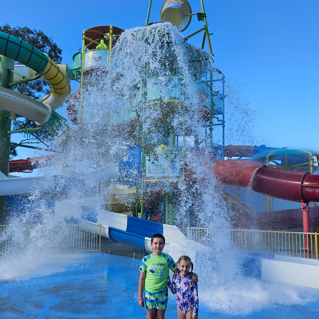 Two Children in Bright Swimsuits Stand Together in a Splash Zone As Water Erupts from a Large Fountain at a Colorful Water Park.