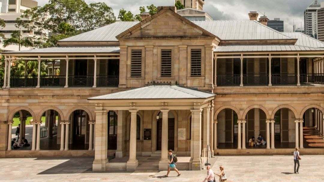 Historic Sandstone Building with Arched Colonnade and Central Portico, People Walking in a Sunny Courtyard.