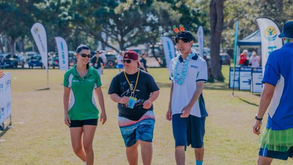 0720_trecforrec_2026_hero | Kids in Brisbane, Gold Coast, Sunshine Coast | Kotc Three People Walk Together at an Outdoor Event: a Woman in a Green Polo, a Man in a Black Shirt and Cap, and a Tall Man in a White Shirt with Leis and Festive Accessories.