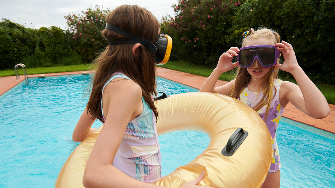 Choose-swimwear-1 | Kids in Brisbane, Gold Coast, Sunshine Coast | Kotc Two Girls by a Pool Wearing Goggles, One Holding a Large Yellow Inflatable Ring Between Them.