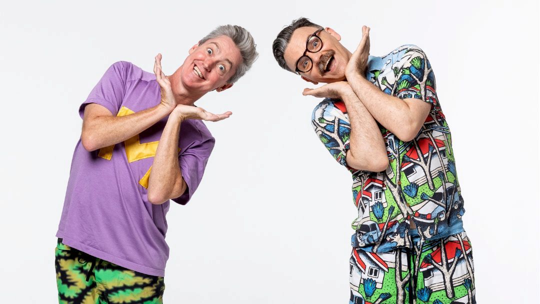 Two Men in Colorful Outfits Pose Playfully, Framing Their Faces with Their Hands Against a White Background.