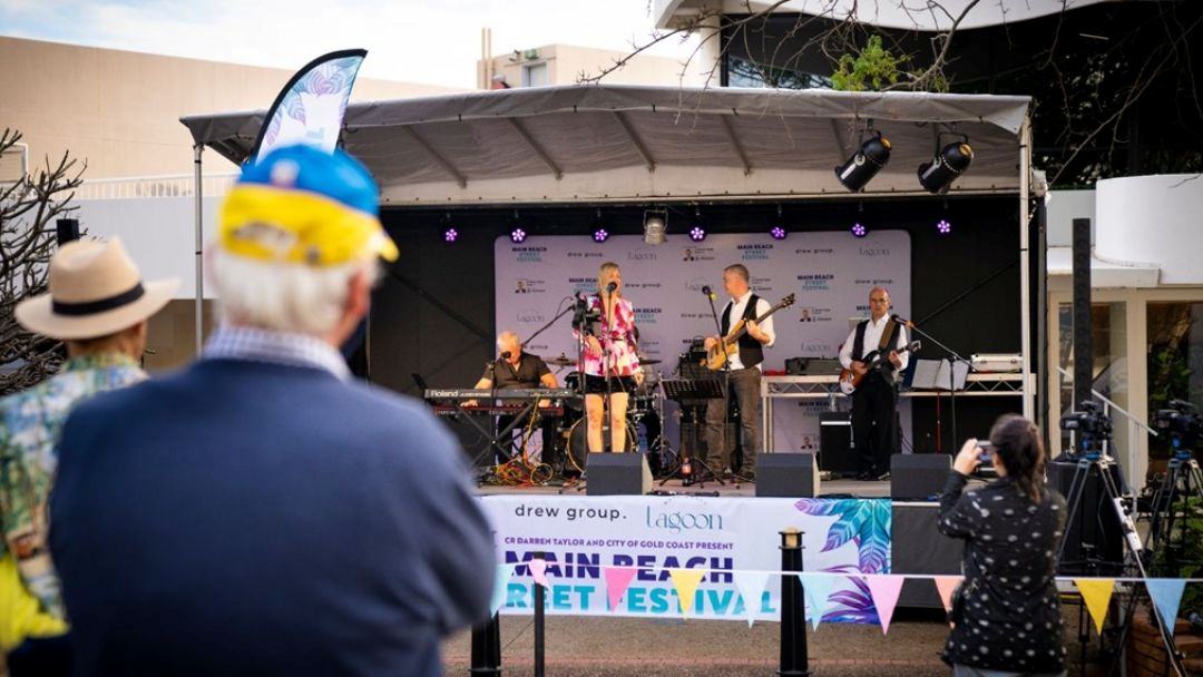 0812_cityofgoldcoast_mainbeachstreetfestival_hero | Kids in Brisbane, Gold Coast, Sunshine Coast | Kotc a Live Band Performs on an Outdoor Stage at a Festival, with a Keyboardist, Guitarist, Bassist and Singer, While Audience Members in the Foreground Watch and Take Photos, and Colorful Bunting Lines the Barrier.