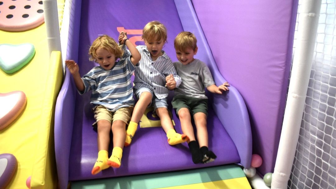 Three young boys cheer as they slide down a purple indoor playground slide, holding hands together.
