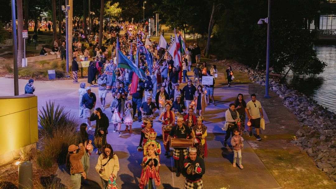 0817_multiculturalaustralia_luminousintheregions_hero | Kids in Brisbane, Gold Coast, Sunshine Coast | Kotc Nighttime Street Parade Along a Waterfront; People Carry Colorful Flags, Drums, and Lanterns.