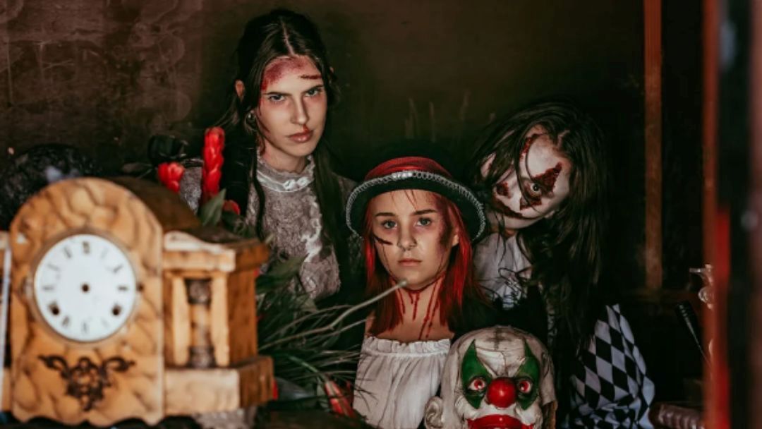 Three Girls with Eerie, Blood-streaked Makeup Pose in a Dark Room, an Antique Clock in the Foreground Adds to the Eerie Scene.