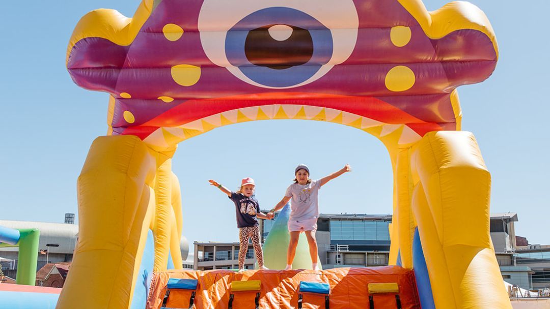 Two Kids Hold Hands and Raise Their Arms on a Bright Inflatable Monster Bounce House Outdoors.