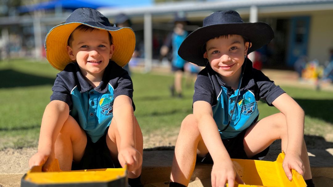Two Smiling Young Boys in Teal Shirts and Wide-brim Hats Crouch on a Yellow Playground Toy Outside a School.