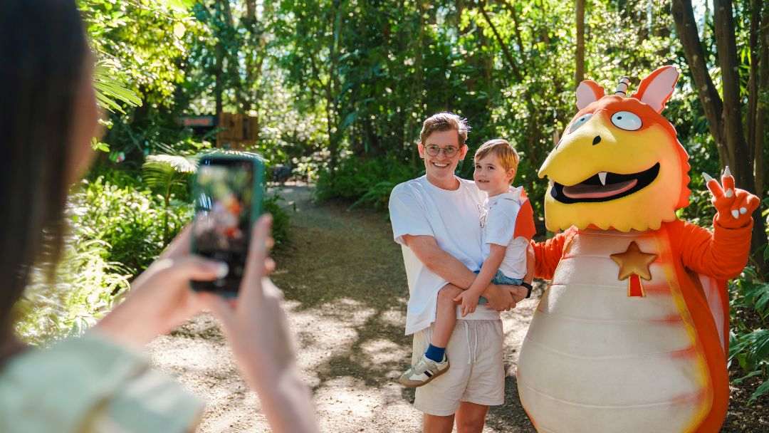 Man and Young Boy Pose with an Orange Dragon Mascot While a Person in the Foreground Takes Their Photo in a Sunny Park Ladscape.