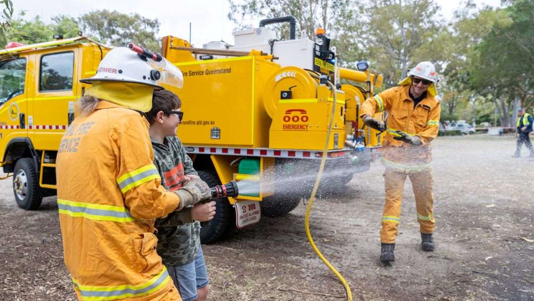 Firefighters in Orange Gear Spray Water from a Yellow Emergency Fire Truck While a Child Helps Hold the Hose.