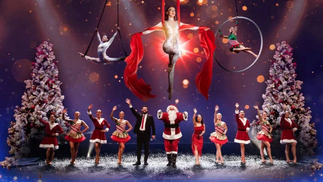 Holiday Stage Show with Dancers in Red Outfits and a Central Santa Figure, Plus Aerial Performers Above a Snowy-looking Floor.