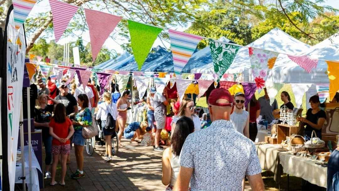 0831_afairdayout-hero | Kids in Brisbane, Gold Coast, Sunshine Coast | Kotc Outdoor Market Scene with Colorful Bunting and People Browsing Vendor Stalls on a Sunny Day.