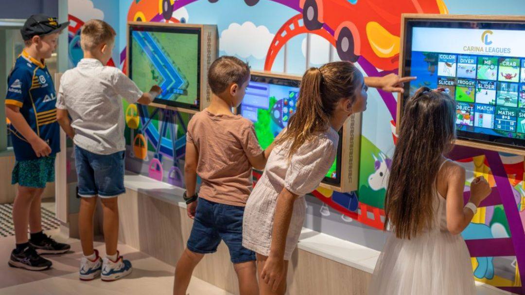 Group of children watching and interacting with colorful touchscreen games in a bright museum exhibit, lively wall art behind them.