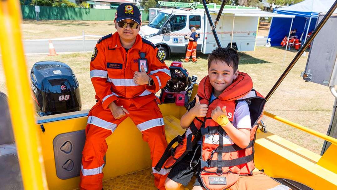 Rescue Worker in Orange Gear and a Boy in a Life Jacket Sit in a Yellow Boat, Giving Thumbs Up; an Ambulance and Tents Are in the Background.
