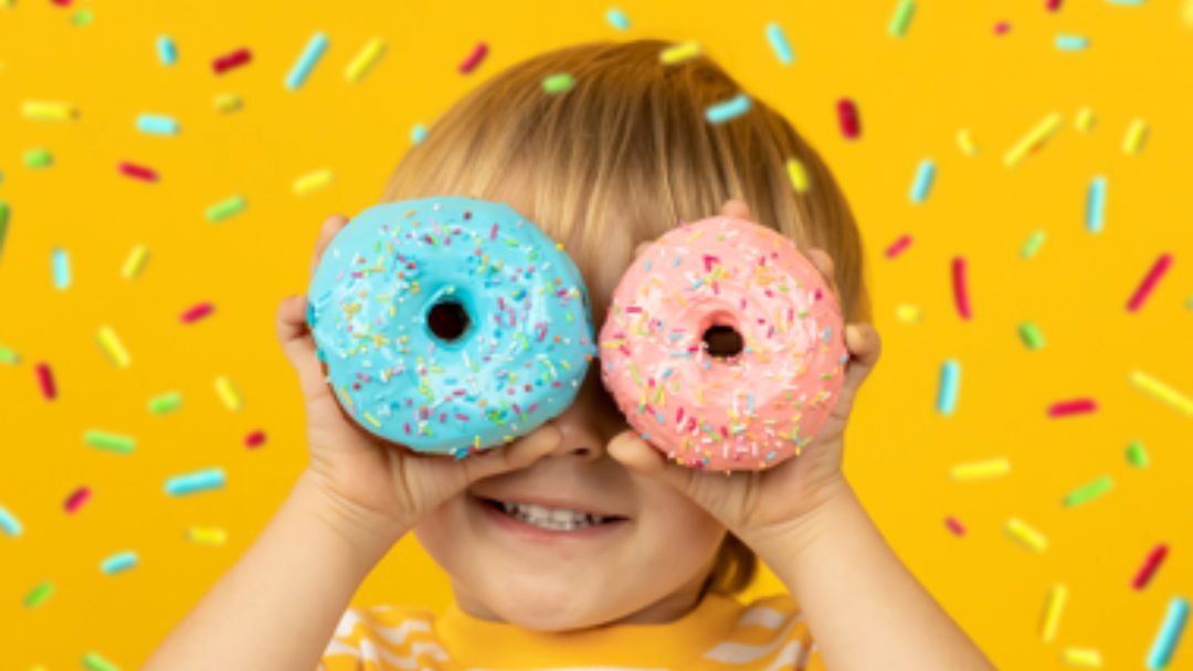 Child Playfully Holds Blue and Pink Sprinkled Donuts over Eyes Against a Bright Yellow Background with Confetti Sprinkles; Smiling Face Visible Beneath.