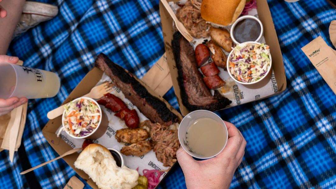 Picnic Spread on a Blue Plaid Blanket Featuring Ribs, Sausage, Chicken, Coleslaw, and Drinks in Disposable Cups.
