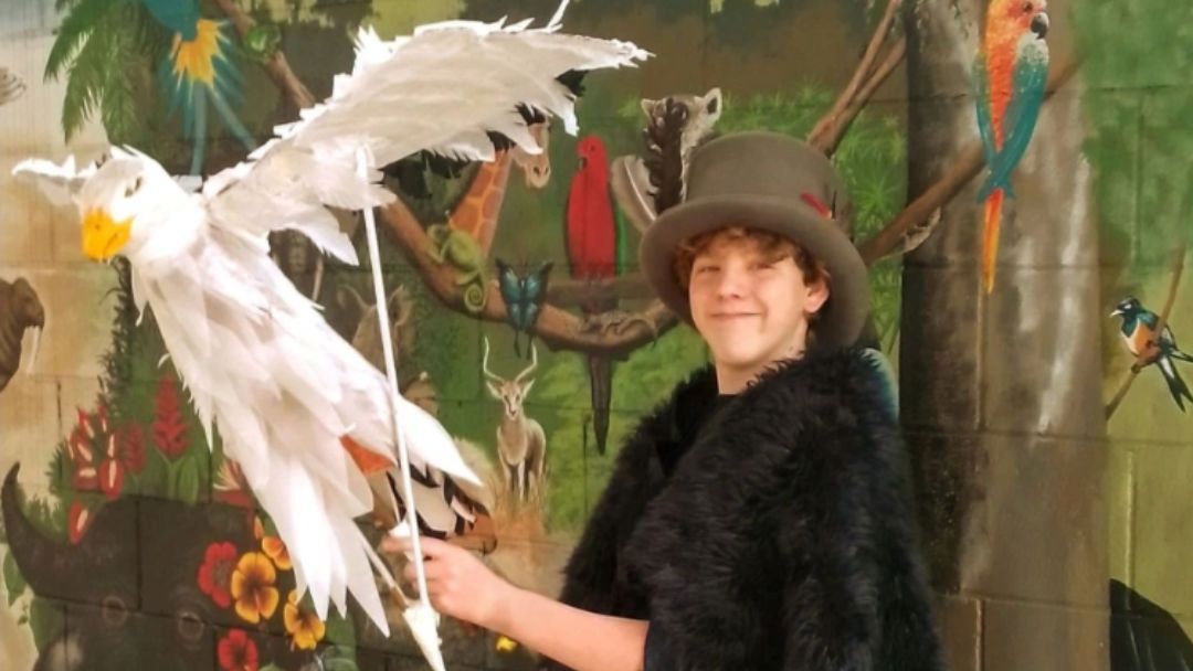0918_thevillagepuppetfestival_hero | Kids in Brisbane, Gold Coast, Sunshine Coast | Kotc Young Person in a Black Fur Coat and Top Hat Posing with a Large White Bird Prop in Front of a Colorful Mural Backdrop