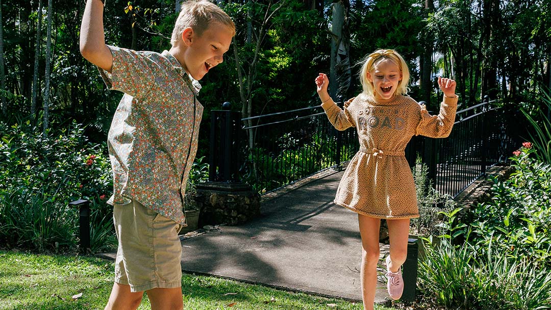 Alsahwa Estate | Kids in Brisbane, Gold Coast, Sunshine Coast | Kotc Two Children Jump with Arms Raised on a Sunny Garden Path, Smiling and Having Fun in a Lush Outdoor Setting.