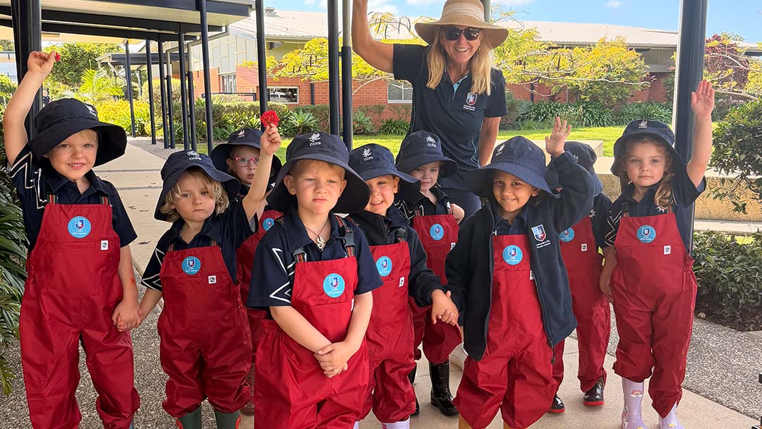 Ccps 1223 1 | Kids in Brisbane, Gold Coast, Sunshine Coast | Kotc Group of Young Children in Red Overalls and Navy Uniforms with Hats, Outdoors with a Smiling Teacher Waving in the Air Behind Them.