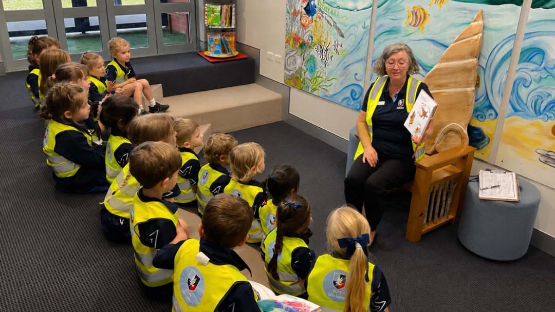 Ccps Classroom Storytime 1 | Kids in Brisbane, Gold Coast, Sunshine Coast | Kotc a Group of Young Children in Bright Safety Vests Sit on the Floor, Listening to a Teacher Reading a Book in a Colorful Mural-filled Room.
