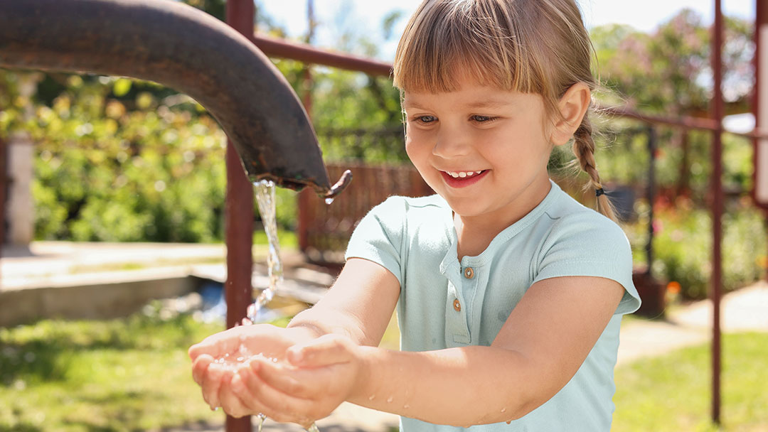 Young Girl at a Playground Cups Water from a Fountain with a Smile, Sunlit Greenery in the Background.