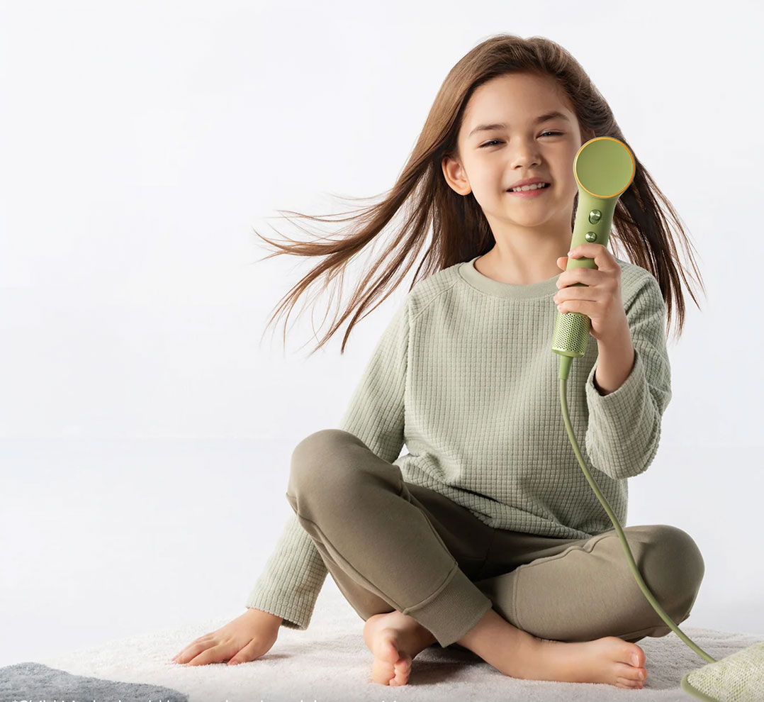 Cto-hairdryer | Kids in Brisbane, Gold Coast, Sunshine Coast | Kotc Young Girl Sitting Cross-legged on a Rug, Smiling, While Holding a Green Handheld Hair Styling Tool with a Circular Head.