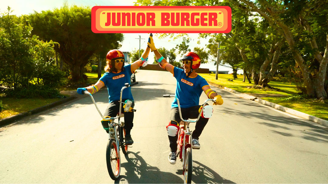 Cto-junior-burger-album | Kids in Brisbane, Gold Coast, Sunshine Coast | Kotc Two People on Bikes Wearing Helmets High-five While Riding Down a Sunny Street; a Junior Burger Sign is Overhead.