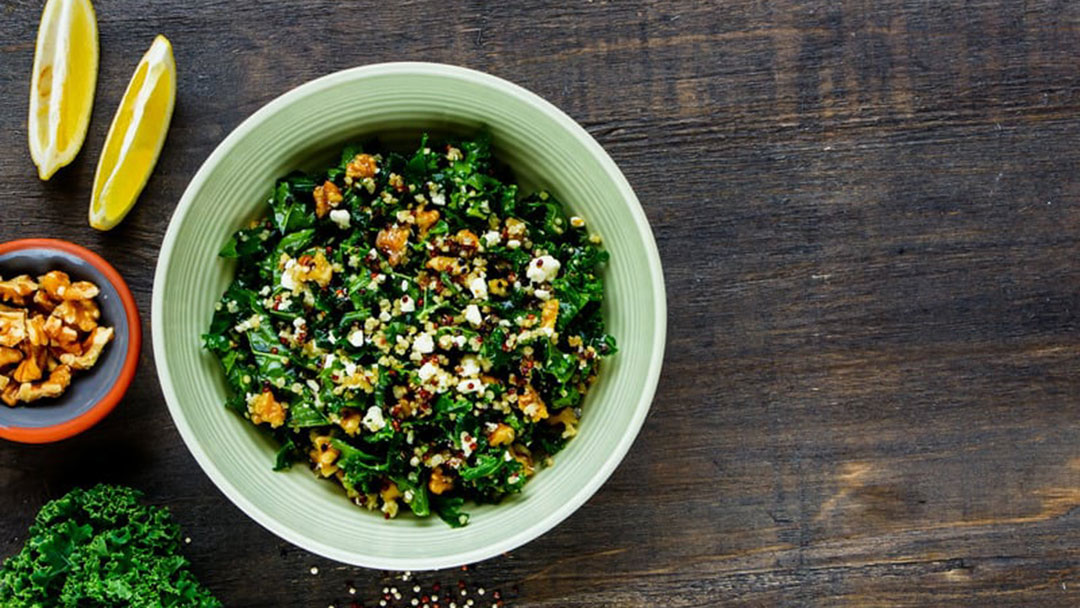 Cto-livinghealthy | Kids in Brisbane, Gold Coast, Sunshine Coast | Kotc Bowl of Kale Salad Topped with Crumbled Cheese and Nuts on a Dark Wooden Table, with Lemon Wedges Nearby.