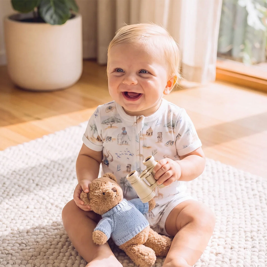 Cto-moobaba | Kids in Brisbane, Gold Coast, Sunshine Coast | Kotc Smiling Baby Sits on a Light Rug Indoors, Holding a Teddy Bear and a Small Pair of Toy Binoculars.