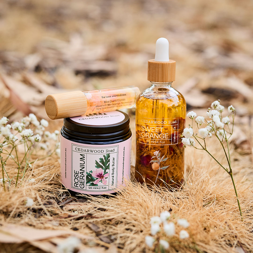Cto-skincare | Kids in Brisbane, Gold Coast, Sunshine Coast | Kotc Cedarwood Road Skincare Set Outdoors: Rose Geranium Body Butter Jar, Sweet Orange Oil with Dropper, and a Small Roller Bottle on Dry Grass with White Flowers.