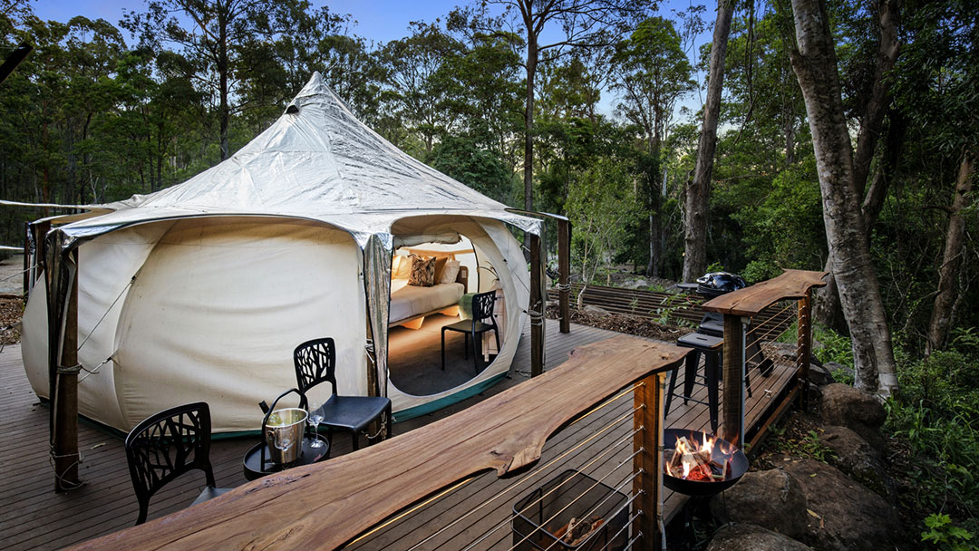 Dig-it-gold-coast-thunderbird-glamping | Kids in Brisbane, Gold Coast, Sunshine Coast | Kotc Glamping Bell Tent on a Wooden Deck in a Forest, Open Doorway Reveals a Bed Inside with Seating and a Nearby Fire Pit on the Deck.
