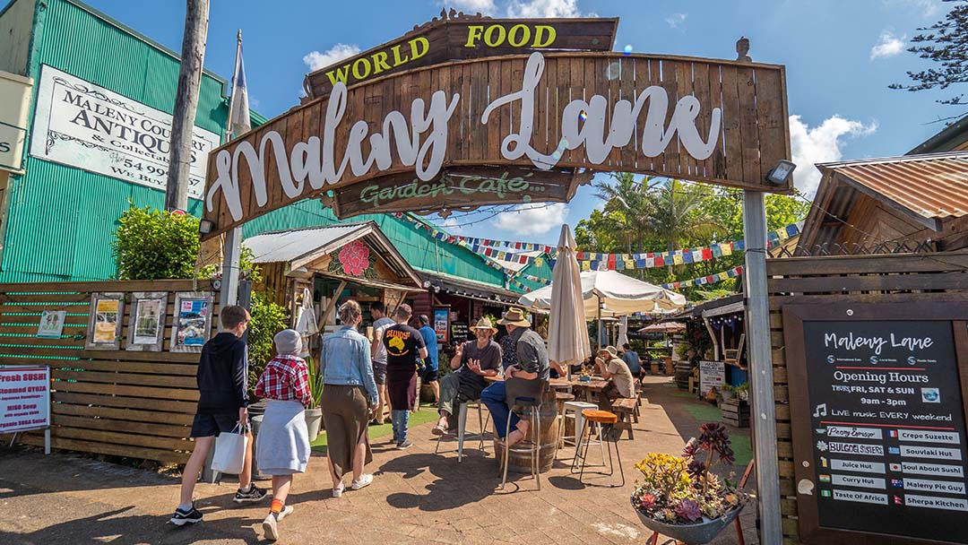 Archway Sign Reading'Maleny Lane' over an outdoor market with people strolling, wooden stalls, and colorful bunting under bright skies.